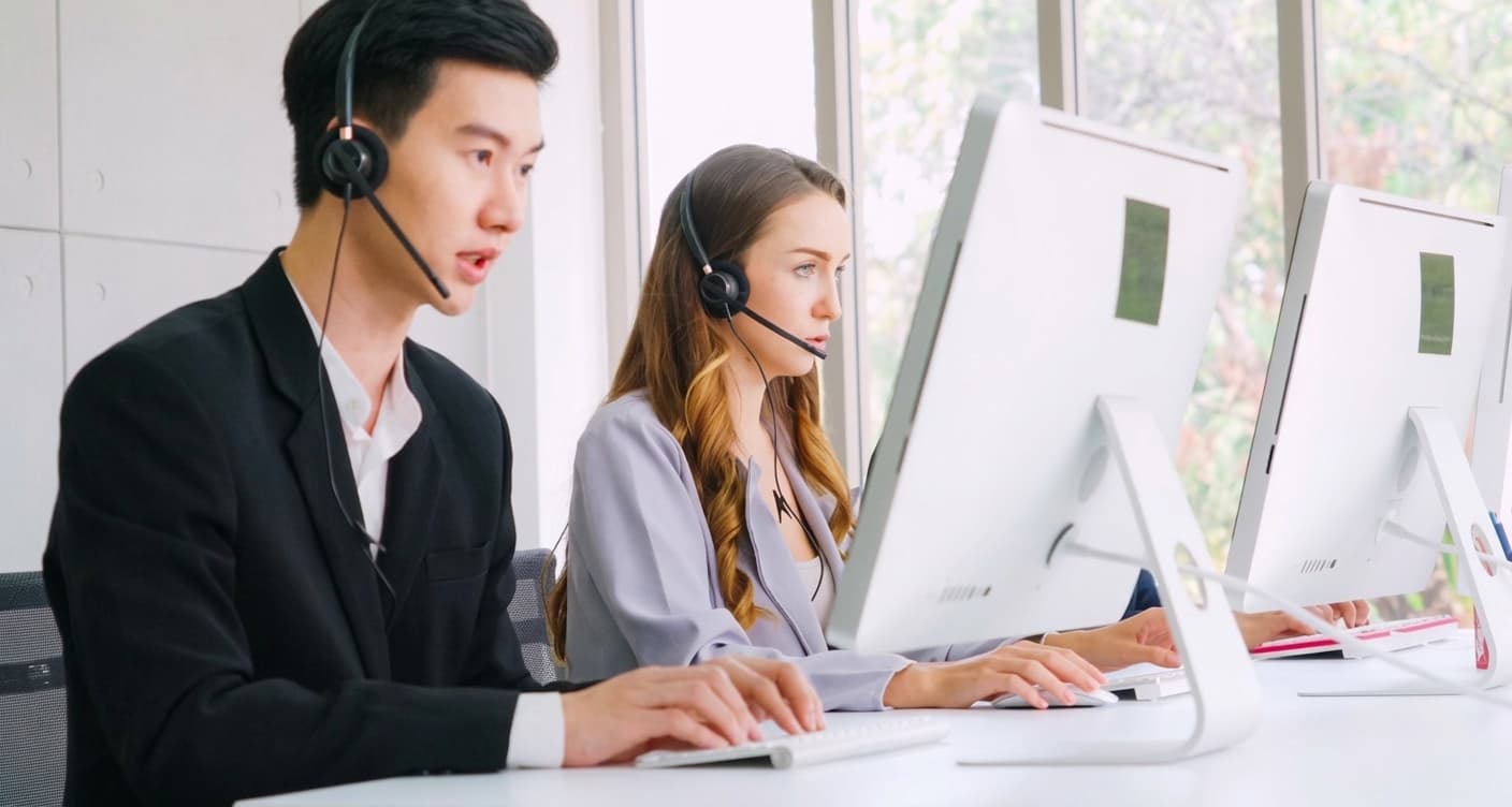 Two agents in headsets talking at desktop computers in a bright office, with no desk phones on the bench