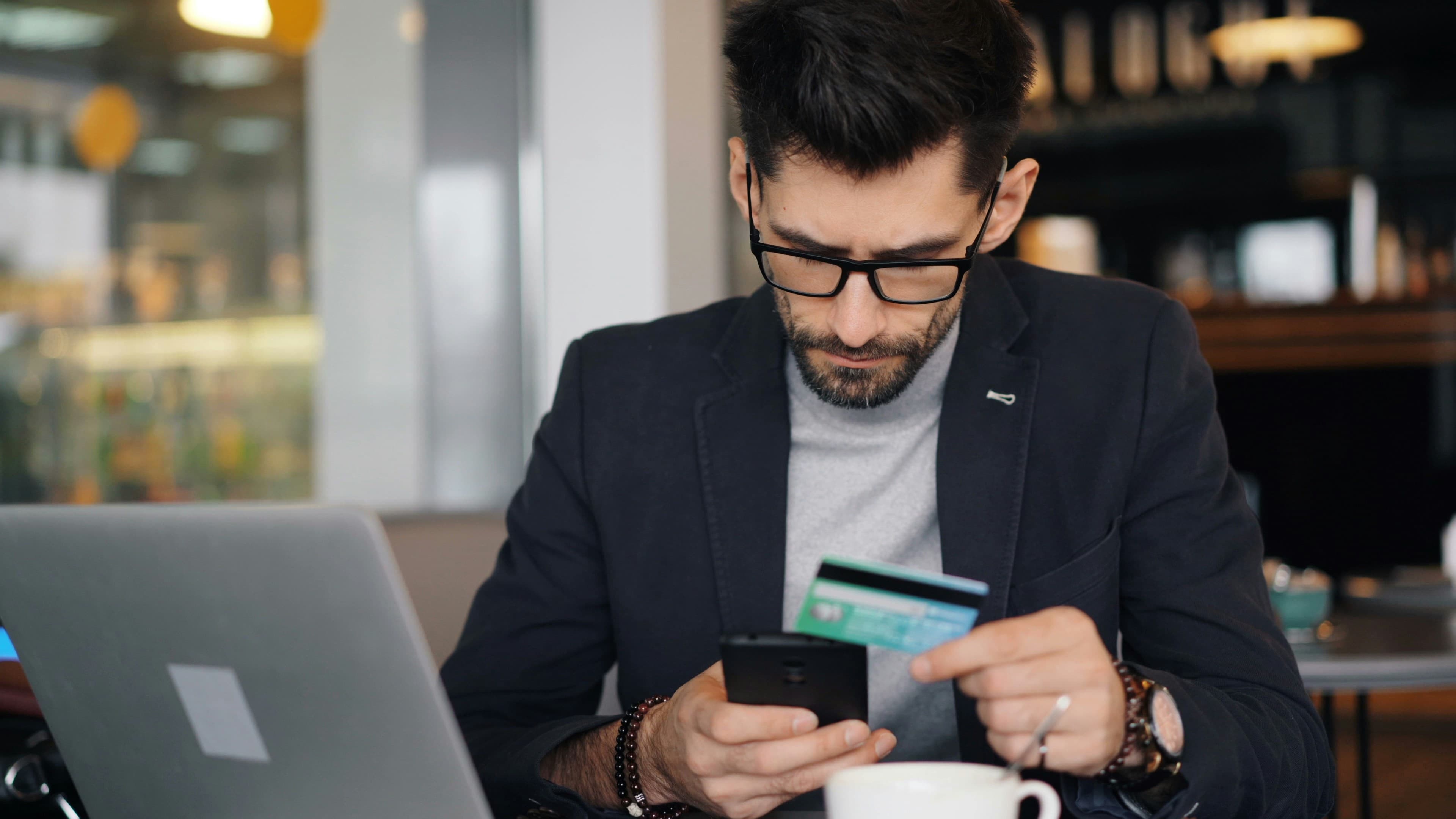 A man at a cafe laptop holding a bank card against his phone, working out what something costs